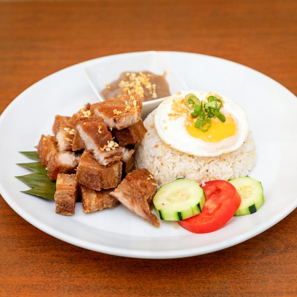 Filipino crispy pork belly (lechon kawali) with rice, fried egg, sauce, and vegetables on a white plate.